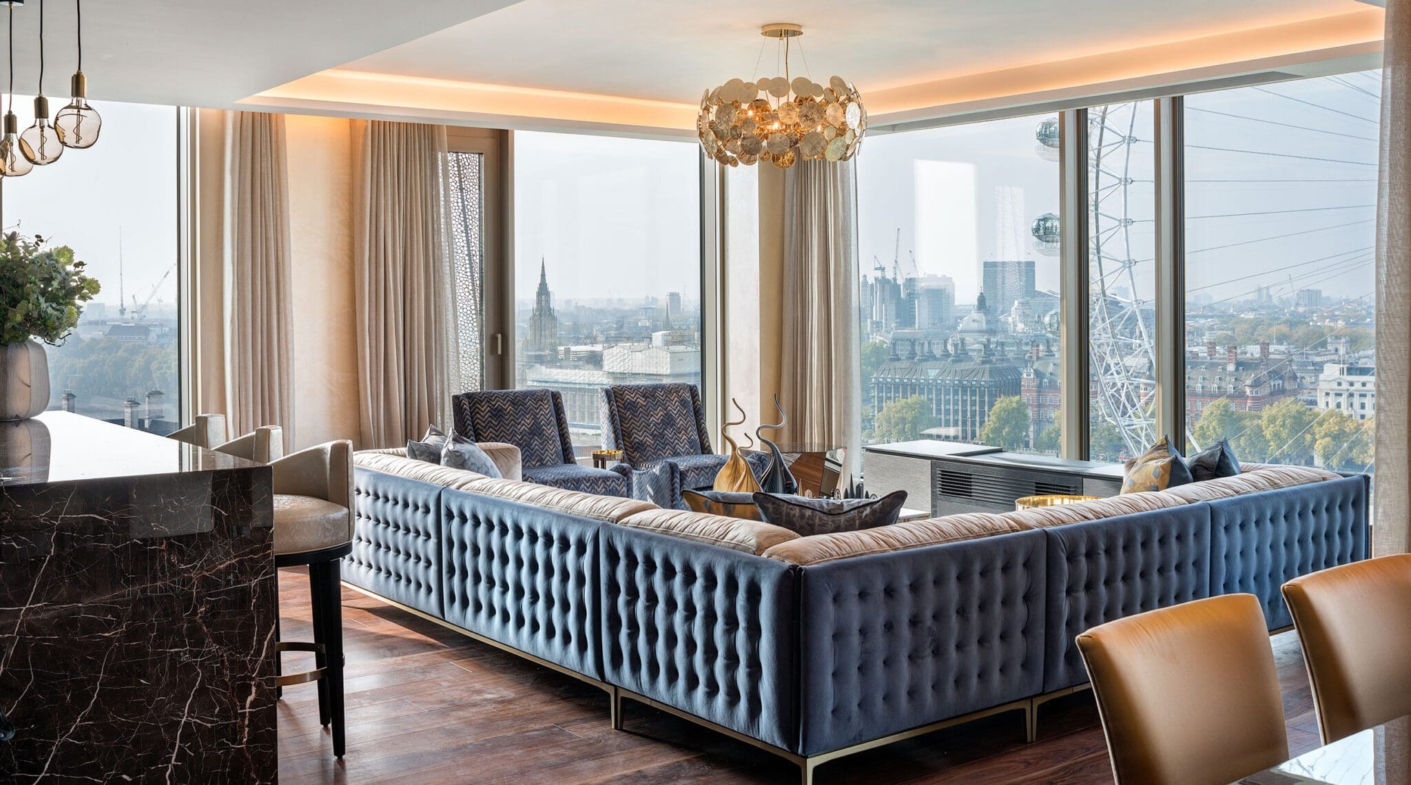 A modern living room showcases a wide image of luxury with a large blue sectional sofa and a marble table. Floor-to-ceiling windows reveal city views, including the London Eye, set against a hazy skyline, capturing the essence of contemporary elegance.