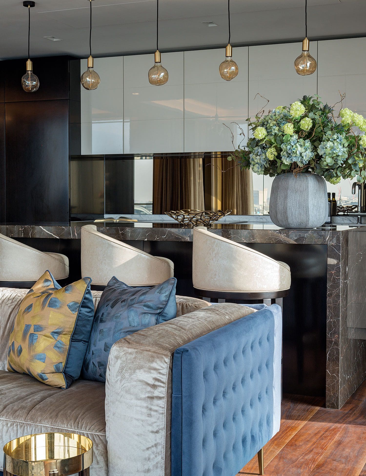 Modern living room from row 08a and b featuring a plush blue and beige sofa, decorative pillows, a marble counter, bar stools, pendant lights, and a large flower arrangement in the background.