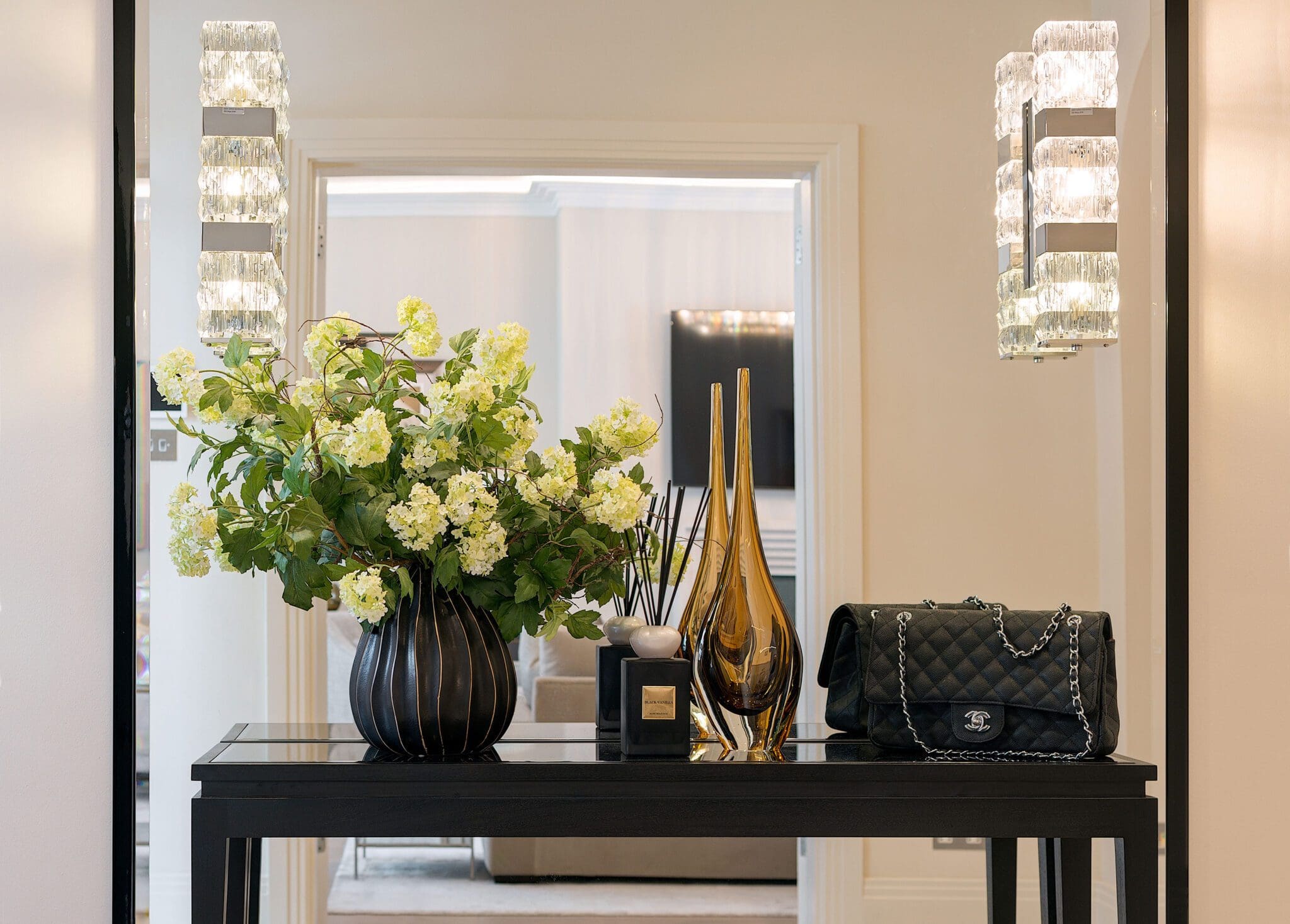 A decorative entry table with a black vase holding white flowers, two amber glass vases, a luxury handbag, and two wall-mounted light fixtures.