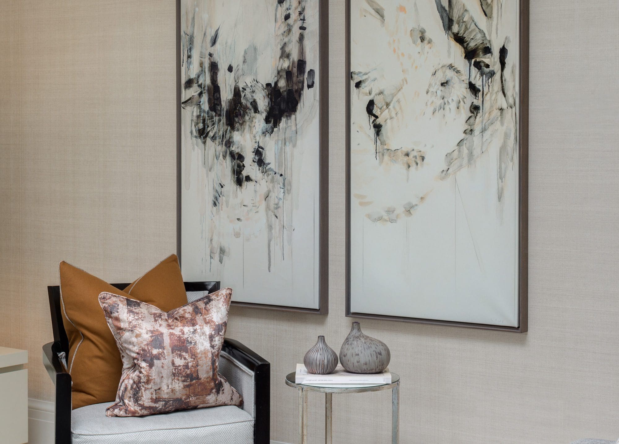 A chair with a patterned pillow is next to a small round table holding two decorative vases. Two abstract black and white paintings are on the beige wall.