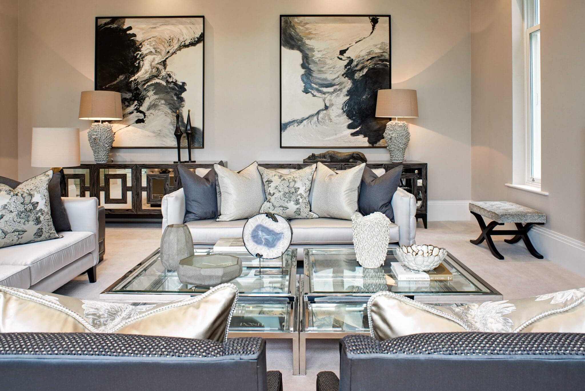 A contemporary living room features two plush white sofas and a sleek glass coffee table, accented with decorative pillows. Abstract art adorns the walls like a header template, while lamps and chic decor items are elegantly arranged on a stylish console table, creating an inviting atmosphere.