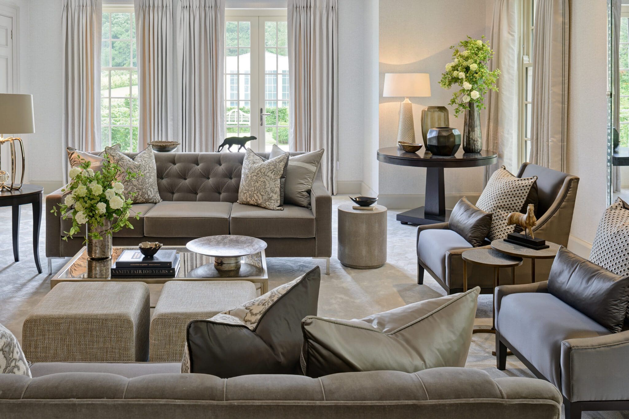 A modern living room with grey sofas, armchairs, and ottomans features large windows and neutral-tone curtains. A vase with flowers sits on a round table adorned with decorative items. The design sets a perfect header template for showcasing contemporary elegance.