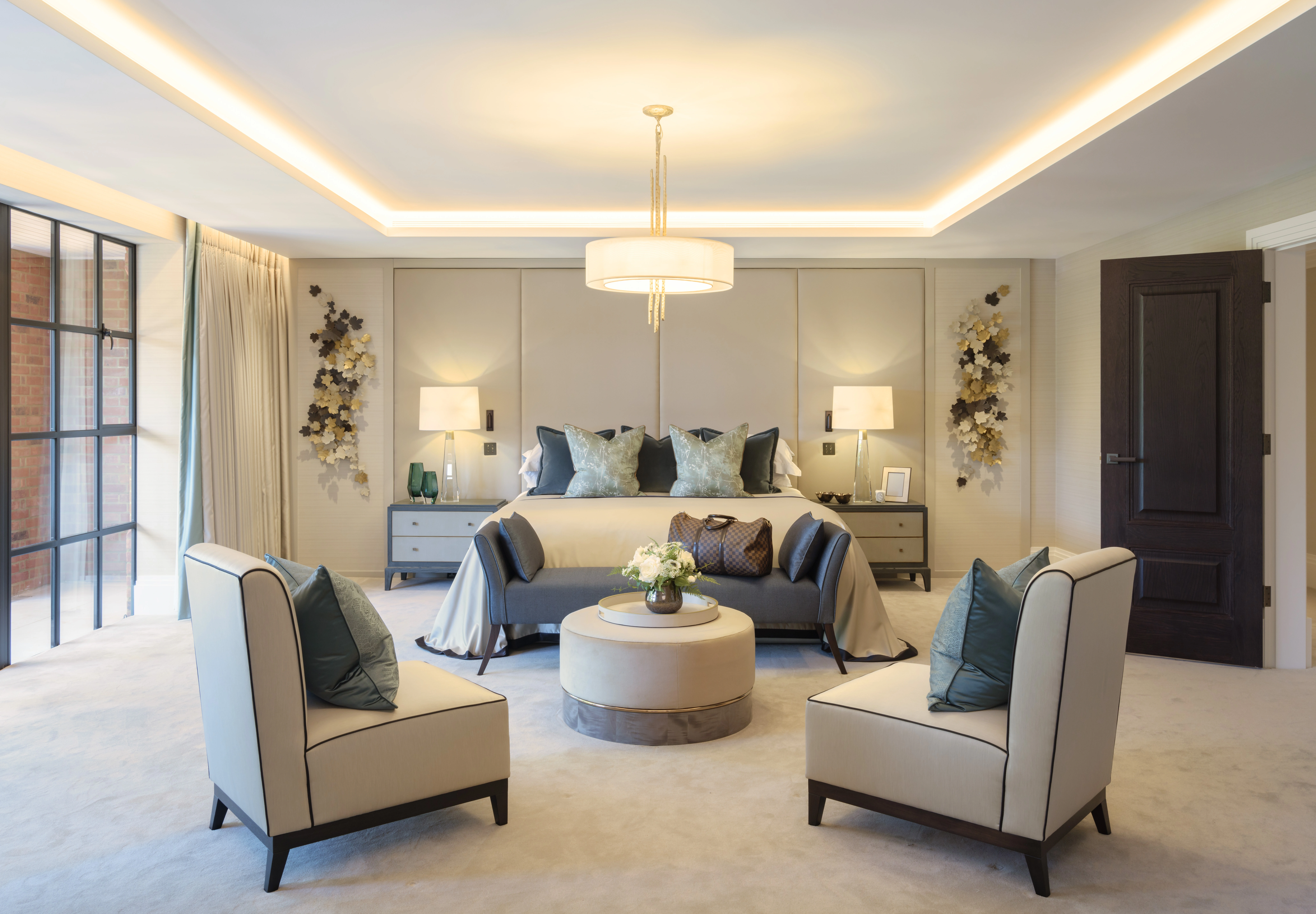 A modern bedroom with a central bed, two chairs, a round ottoman, and decorative lighting. Cream and gray tones with accent pillows and floral wall art create an elegant atmosphere.