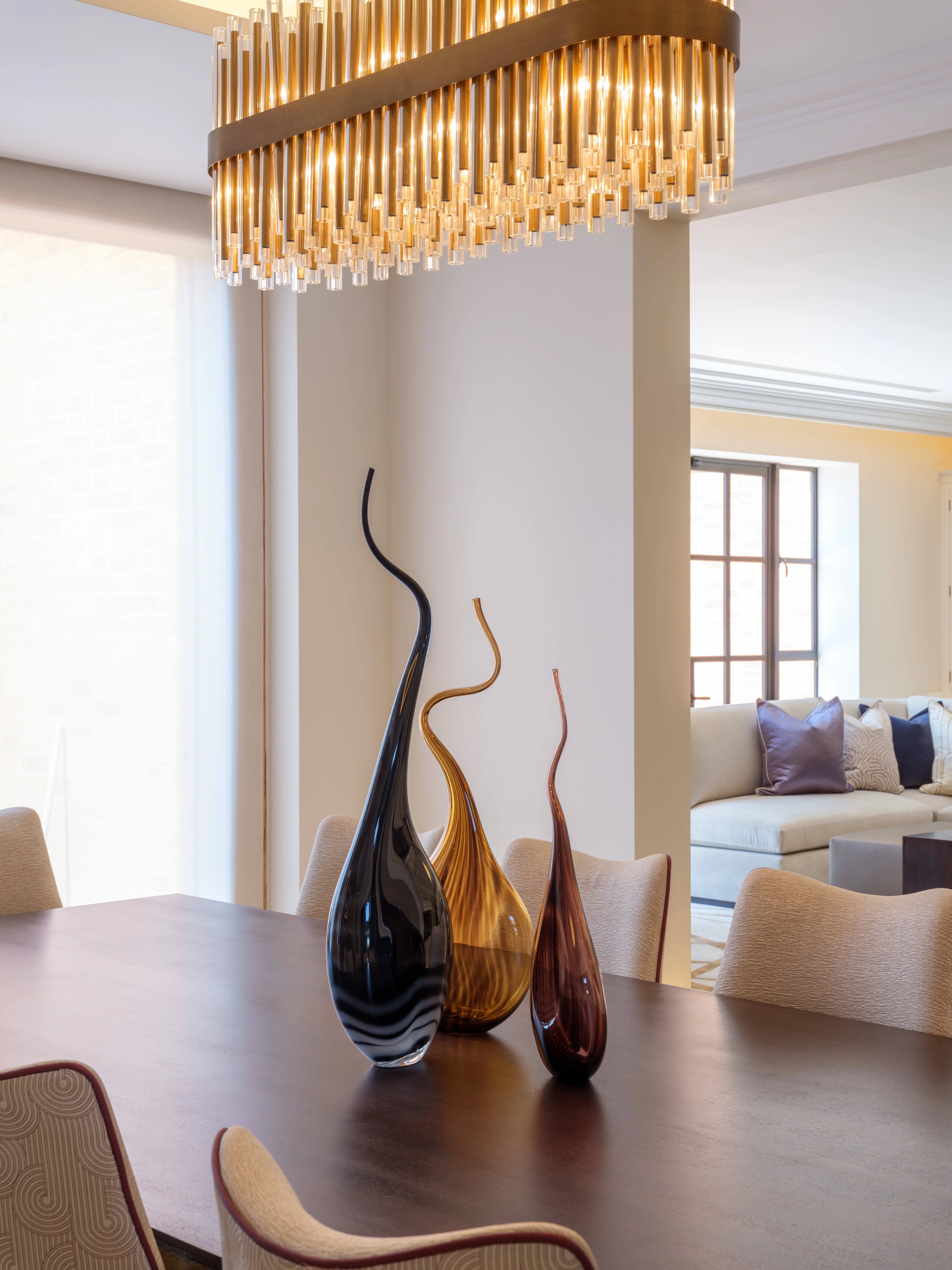 A modern dining room with a dark wood table, sculptural vases, and a contemporary chandelier. A sofa and large windows are visible in the background.