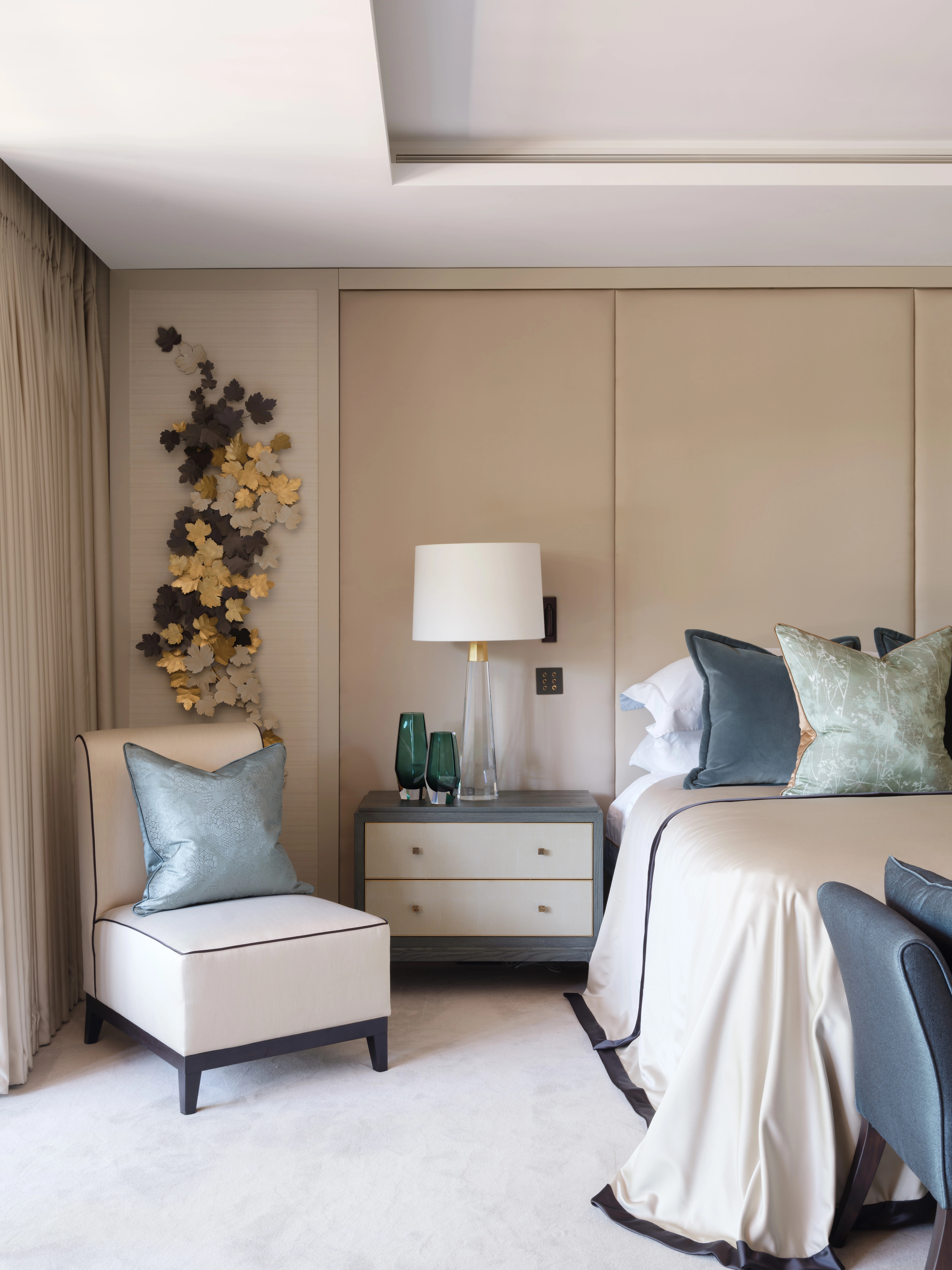 Elegant bedroom with a bed, decorative pillows, a beige chair, and a nightstand holding a lamp and green glass vases. Wall features metallic leaf art. Neutral tones throughout.